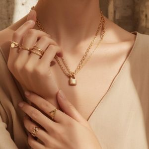 Woman wearing trendy gold-plated jewelry set featuring hoop earrings, layered chain necklace with lock pendant, statement rings, and elegant bracelets in a minimalist fashion style.